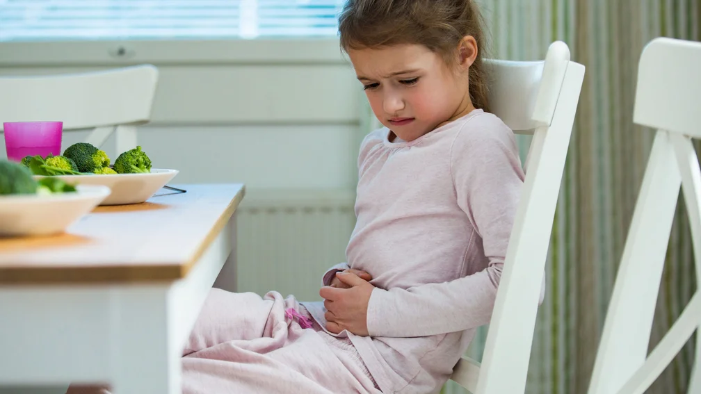 Mädchen sitzt am Tisch, vor ihr steht Essen und sie fasst sich an den Bauch mit einem schmerzverzerrten Gesicht