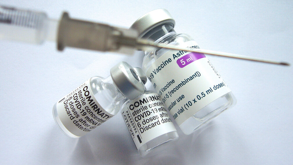 Verschwommen im Vordergrund eine Kanüle, darunter 2 Vial Comirnaty und 1 Vial Astrazeneca