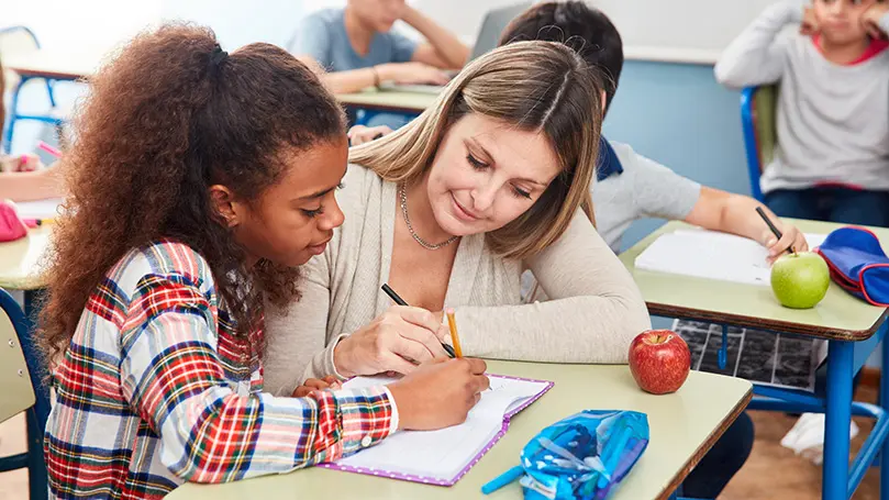Lehrerin kniet an Tisch einer Schülerin und deutet mit Stift auf ein Heft
