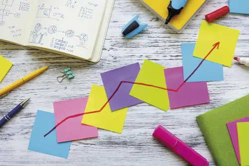 acht bunte Post-its kleben auf einem Schreibtisch aneinander. Darauf zieht sich ein roter Pfeil nach oben durch alle Post-its.