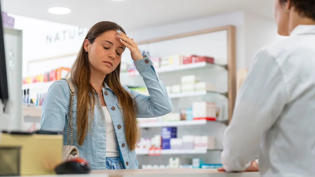 junge Frau steht in der Apotheke und fasst sich mit der Hand an den Kopf