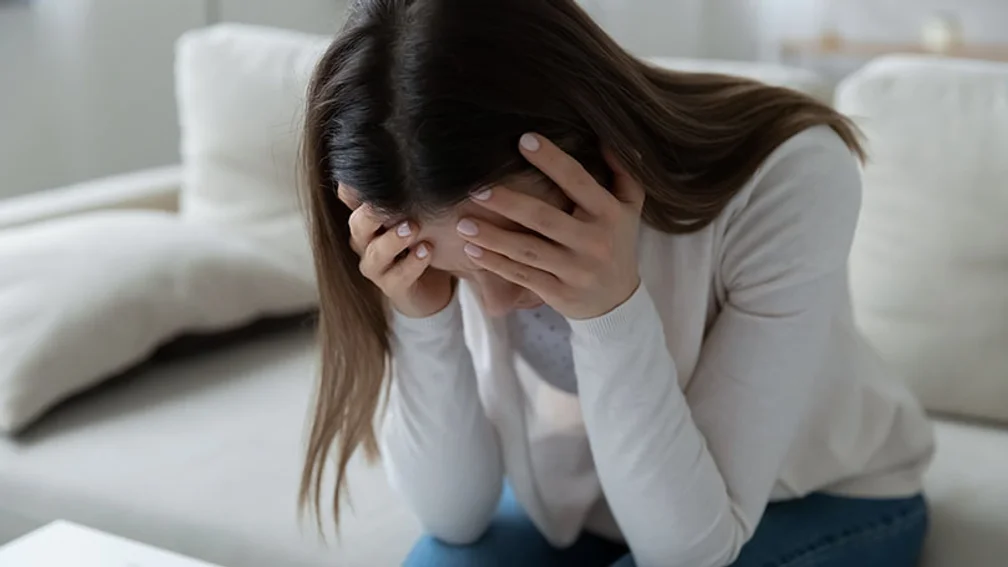 junge, dunkelhaarige Frau sitzt mit in die Hände gestütztem Kopf auf einem weißen Sofa