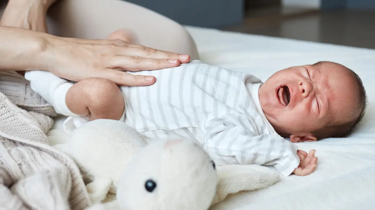 Baby liegt auf dem Rücken und schreit; Hand der Mutter liegt auf dem Babybauch