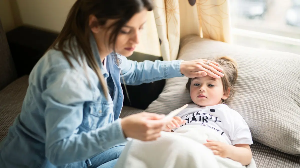 Mutter fasst die Stirn ihrer Tochter mit de rHand an und hält in der anderen Hand ein Fieberthermometer