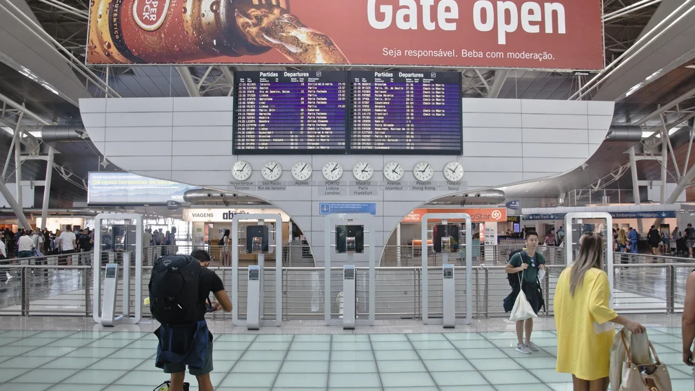 Menschen stehen vor der Fluganzeigetafel, darunter hängen Uhren mit verschiedenen Uhrzeiten, je nach Ort