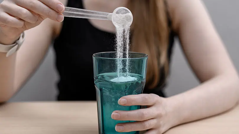 junge Frau schüttet weißes Pulver aus einem Messlöffel in ein grünes Glas mit Wasser