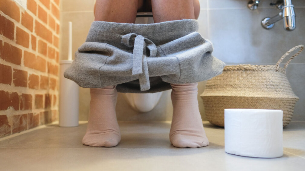 Frau sitzt auf Toilette, Toilettenpapier liegt daneben auf dem Boden