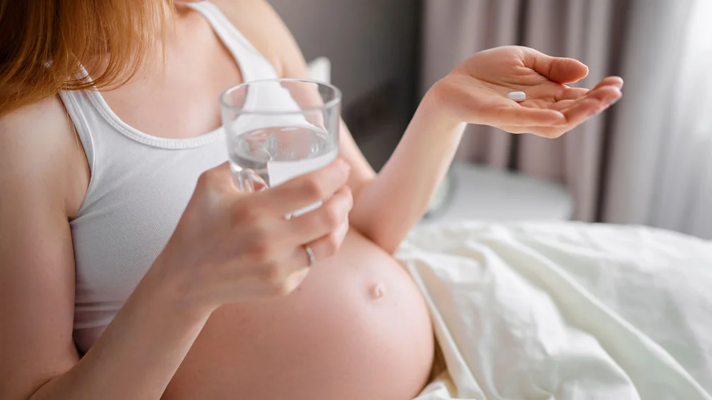 Schwangere liegt im Bett und hält eine Tablette in der einen Hand und ein Glas Wasser in der anderen 