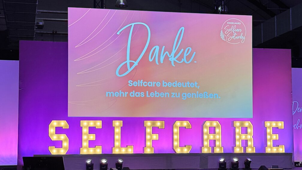 Auf einer Leinwand steht das Wort Danke, darunter große Leuchtbuchstaben, die das Wort Selfcare bilden