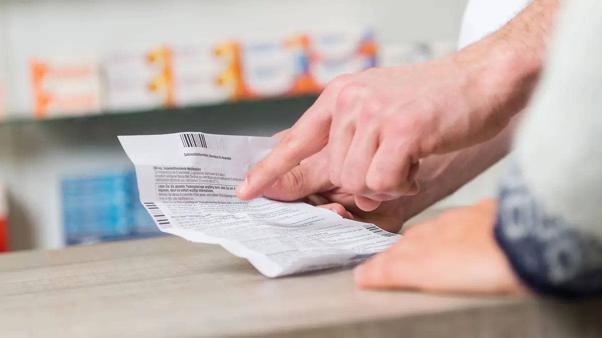 Beipackzettel wird in de rHand eines PTA gehalten, daneben ist eine auf dem HV-Tisch abstützende Hand einer Kundin zu sehen 