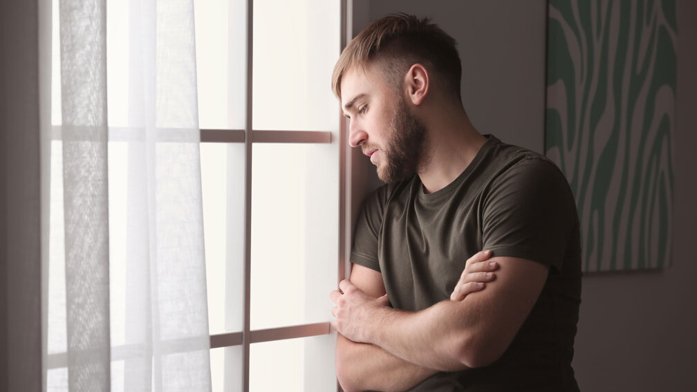 Mann steht vor dem fenster mit einem traurigen Blick und verschränkten Händen vor der Brust