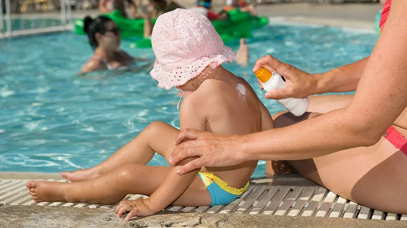 Frau sprüht Sonnencreme auf den Rücken eines Kindes mit Sonnenhut, das am Rand eines Schwimmbecken sitzt
