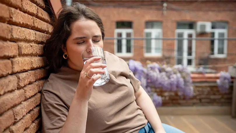 junge geschwächte Frau sitzt an einer Backsteinmauer lehnend und trinkt mit geschlossenen Augen aus einem Glas Wasser
