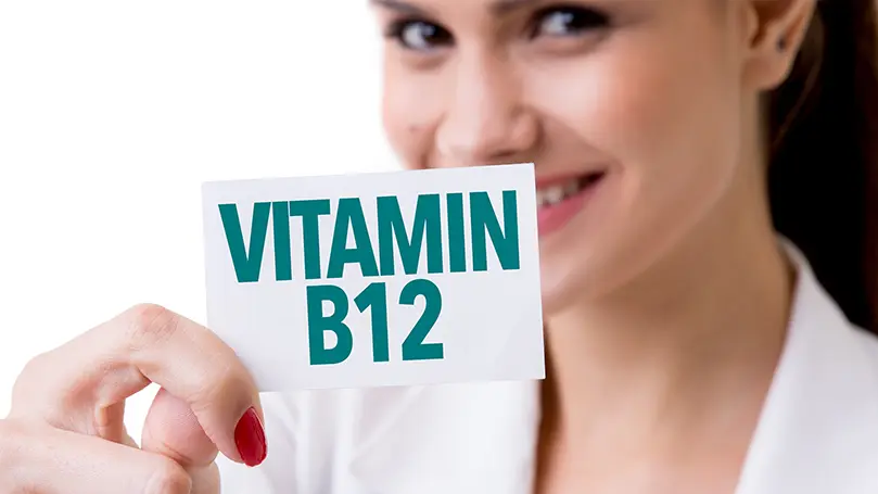 Junge Frau in weißem Arztkittel hält Karteikarte mit den Worten "Vitamin B12" hoch