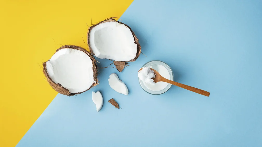 Eine aufgeschnittene Kokosnuss, daneben eine Schale mit Kokosöl und einem Holzlöffel darauf