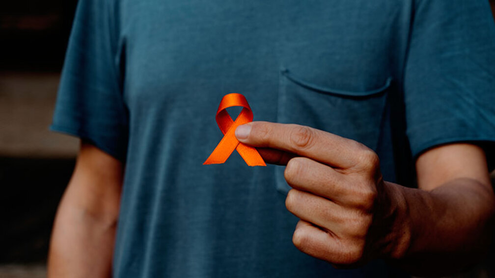 Mann im blauen Tshirt hält orangefarbene Schleife zwischen zwei Fingern