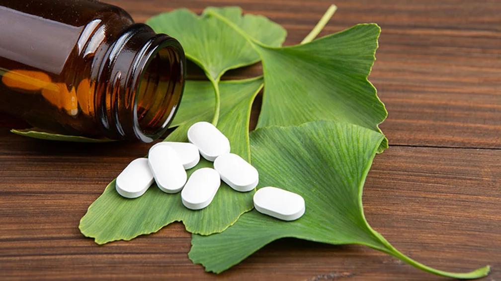 weiße Tabletten liegen auf Ginkgo-Blättern neben einer offenen Braunglasdose