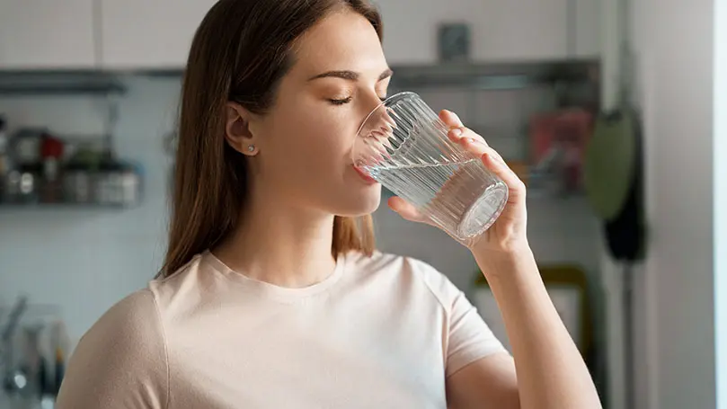 junge dunkelhaarige Frau steht in einer Küche und trinkt mit geschlossenen Augen Wasser aus einem Glas