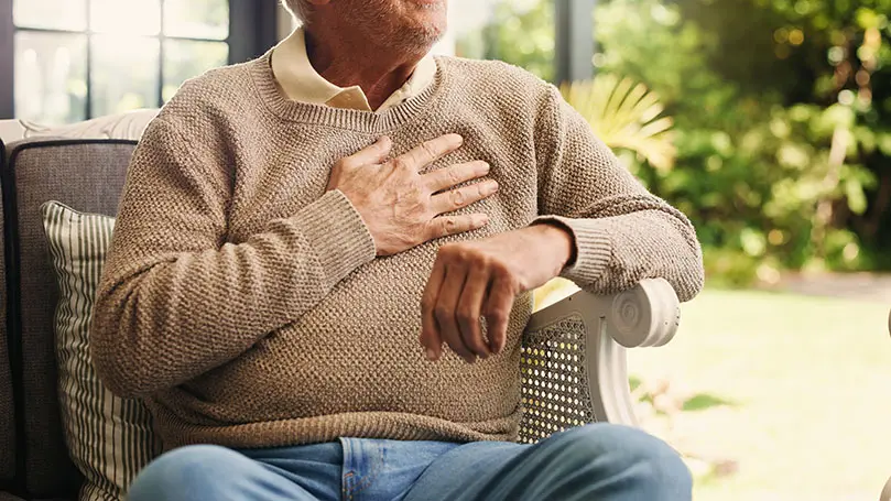 Senior sitzt in einem Rattanstuhl und hat rechte Hand auf sein Herz gelegt