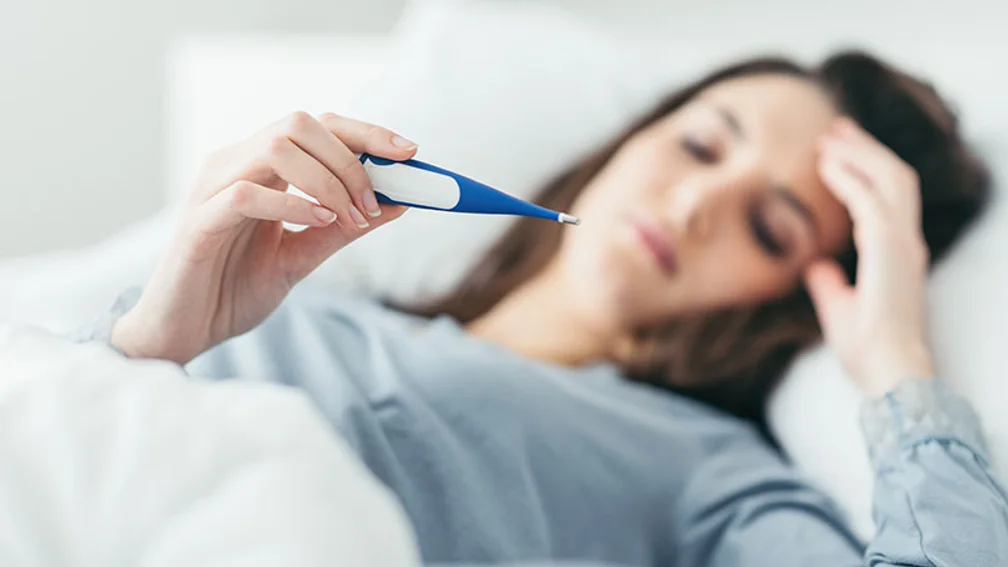 Im Bett liegende Frau blickt auf ein Fieberthermometer
