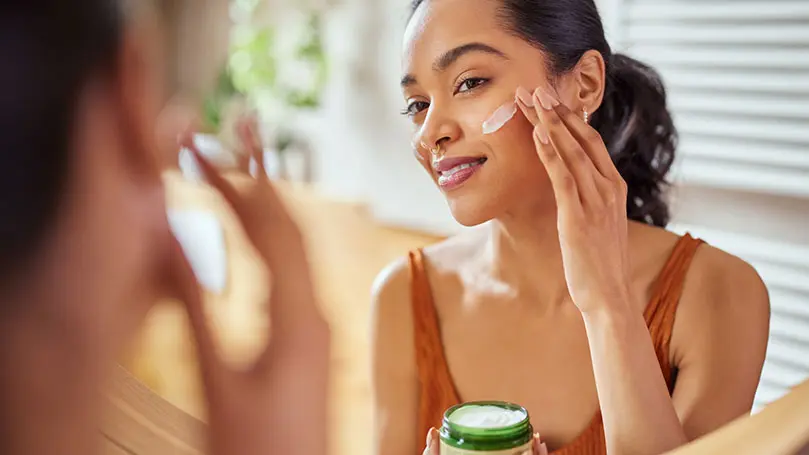 junge dunkelhäutige Frau streicht sich vorm Spiegel Creme auf die Wange