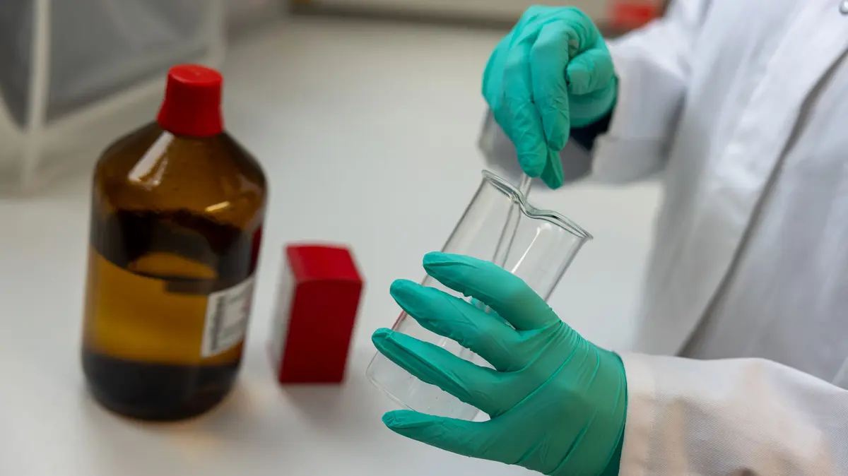 PTA in der Rezeptur und rührt mit einem Glasstab in einem Becherglas, daneben steht ein Braunglasflasche und eine rote Verpackung