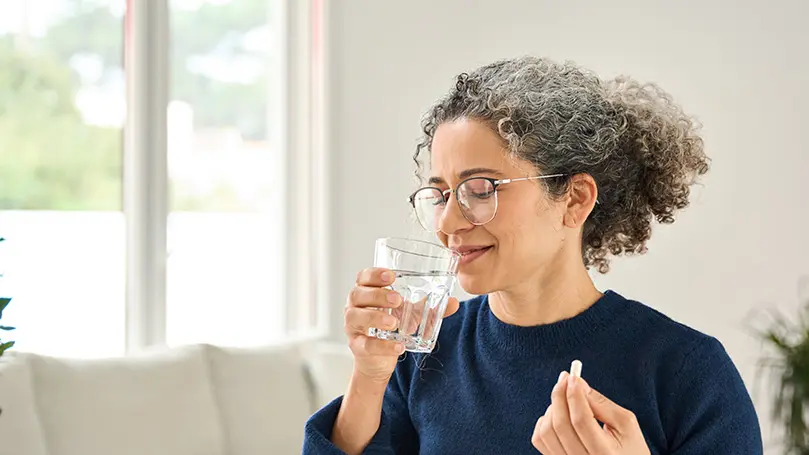 Frau mit leicht ergrautem lockigen Haar trinkt aus einem Wasserglas und hält eine weiße Kapsel in der Hand
