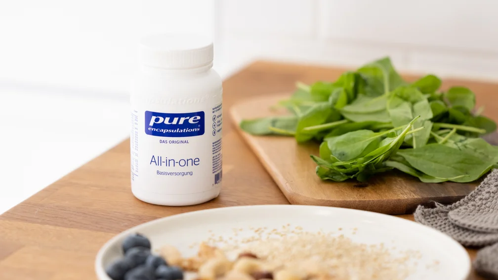 Dose des Nahrungsergänzungsmittels ‚pure encapsulations All-in-one Basisversorgung‘ steht auf einem Holztisch. Daneben liegt frischer Spinat auf einem Schneidebrett. Im Vordergrund ein Frühstücksteller mit Blaubeeren, Nüssen und Haferflocken.