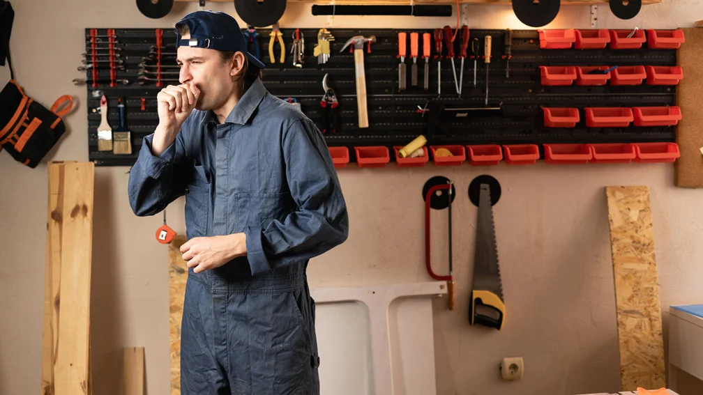 Mann in Arbeitskleidung auf der Arbeit und hustet in die Faust