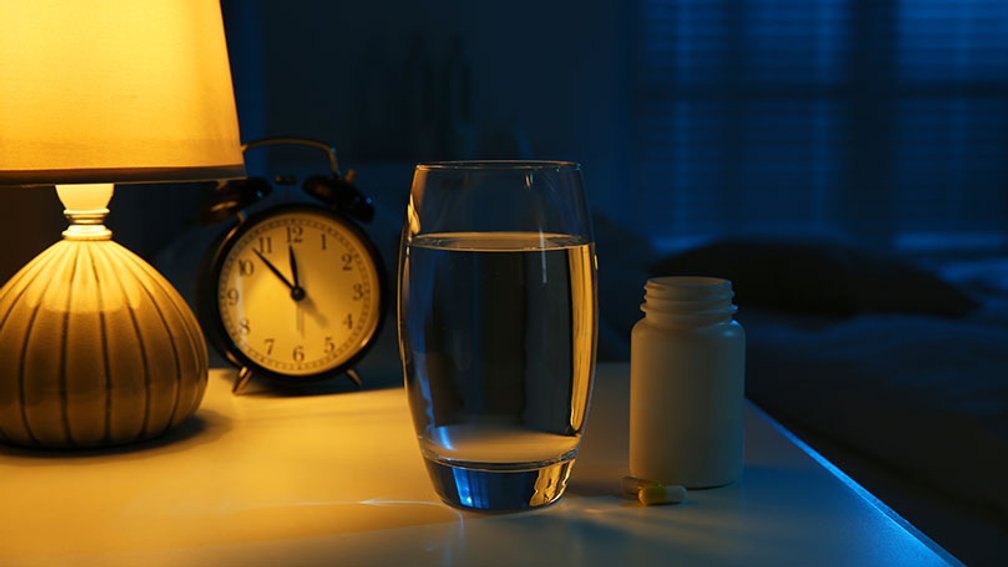 gefülltes Wasserglas und eine Tablettendose stehen auf einem Nachttisch mit Wecker, der kurz vor Mitternacht anzeigt