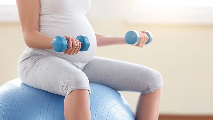 Schwangere in Sportkleidung hält zwei Hanteln in den Händen und sitzt auf einem Gymnastikball