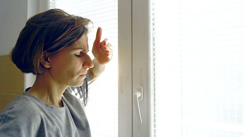 Frau steht vor Fenster mit geschlossenen Augen und hält Hand vor das Gesicht, weil die Sonne sie blendet
