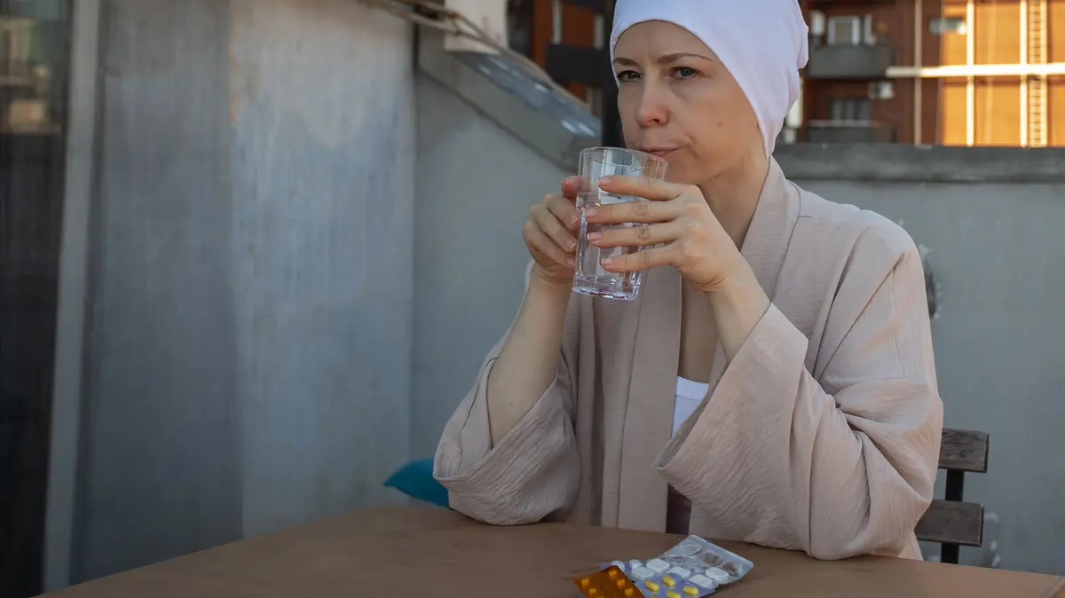 krebskranke Frau hält ein Glas Wasser in der Hand und vor ihr liegen Tablettenblister