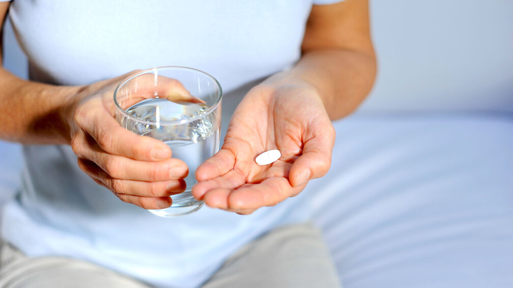 in einer Hand liegt eine weiße Tablette, die andere Hand hält ein Glas Wasser