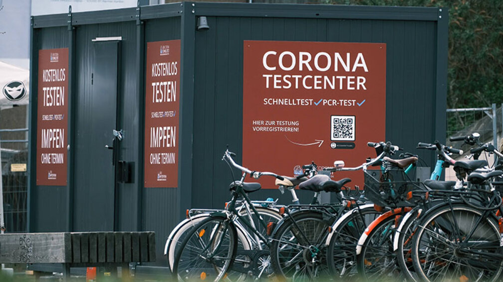 Auf grauem Container klebt rotes Plakat mit Schriftzug Corona Testcenter