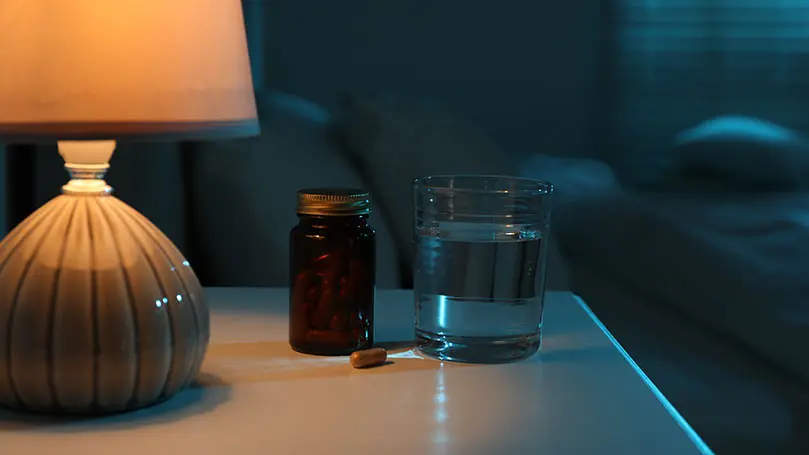 Ein Nachttisch wird von einer Lampe erhellt, darauf steht ein gefülltes Wasserglas neben eine Kapseldose