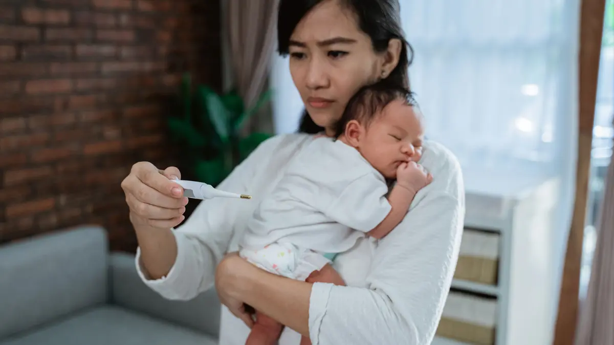 Mutter hält Säugling im arm und schaut auf das Fieberthermometer