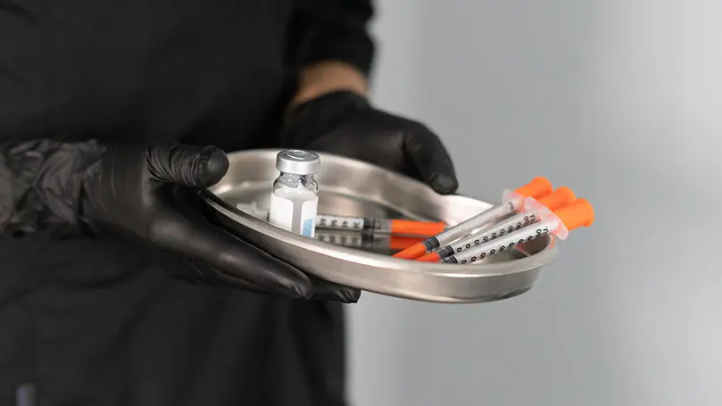 Person mit schwarzen Gummihandschuhen hält Metallschale mit vier Spritzen und einem Impfvial