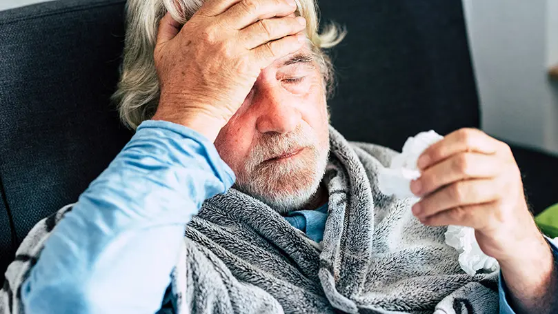 Kranker Senior hält mit geschlossenen Augen eine Hand an der Stirn und in der anderen Hand ein benutztes Taschentuch