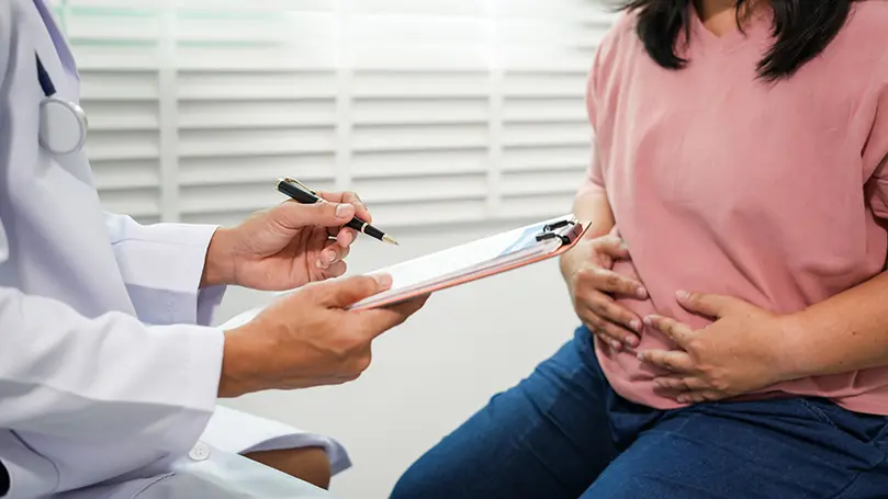 Frau sitzt vor einem Arzt und hält beide Hände an den schmerzenden Bauch