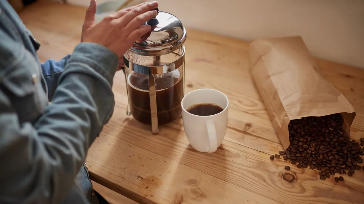 Kaffeebohnen purzeln aus einer Tüte raus, daneben steht eine Person die Kaffee zubereitet