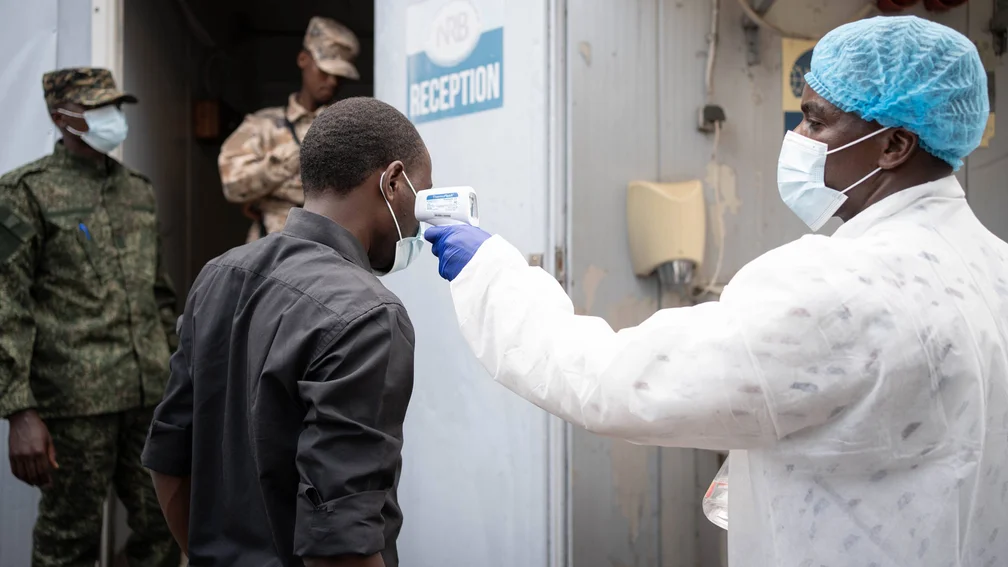 medizinischer Mitarbeiter mit Mundschutz misst die Temperatur eines Anwohners aus dem Kongo; zwei Männer in Uniform stehen im Hintergrund