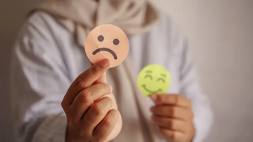Frau hält einen traurigen Smiley in die Kamera, in der anderen Hand hält sie einen lachenden Smiley