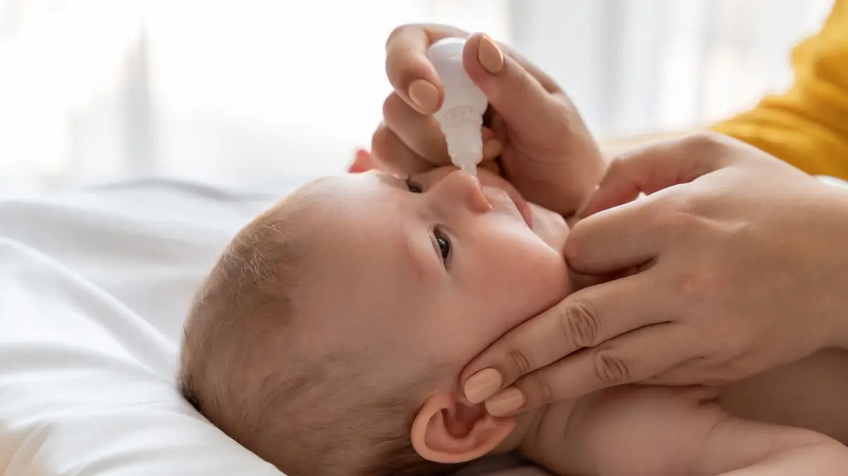 Mutter hält Nasentropfen in der Hand; Säugling liegt vor ihr auf dem Rücken