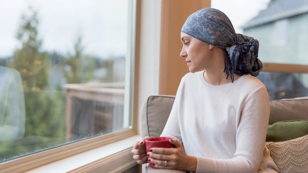 Frau mit Krebserkrankung und Kopftuch hält eine Tasse in den händen und sitzt vor einem Fenster und schaut hinaus