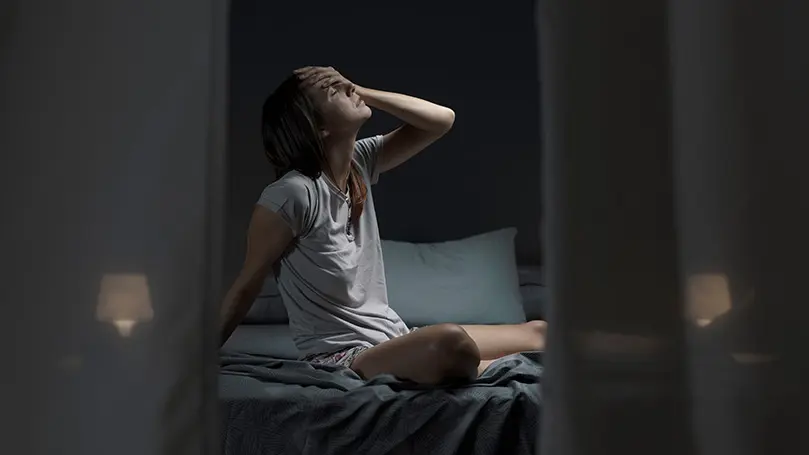 Frau sitzt nachts auf ihrem Bett und hält sich erschöpft die schweißnasse Stirn