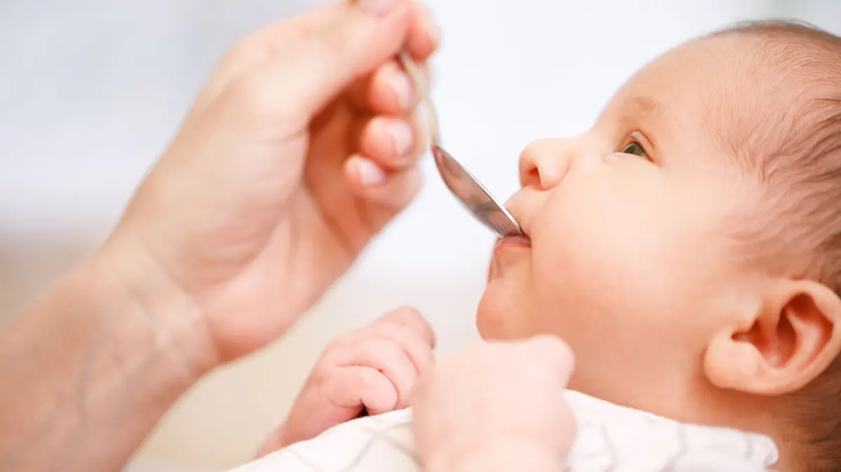 Mutter füttert Säugling mithilfe einem Löffel mit flüssigem Medikament 