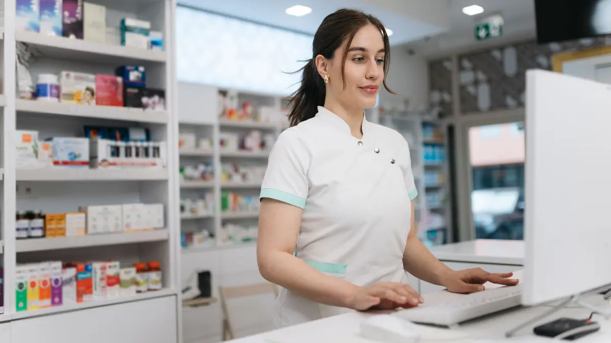 PTA steht in der Apotheke vor dem PC, im Hintergrund sind Regale mit Arzneimittelverpackungen