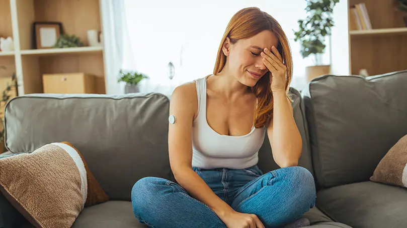 Blonde Frau mit Blutzuckersensor am rechten Oberarm sitzt verzweifelt auf einem Sofa