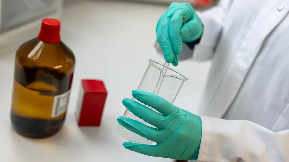 PTA steht in der Rezeptur und rührt mit einen Glasstab in einem Becherglas, daneben steht eine Braunglasflasche mit Flüssigkeit